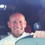 A man smiling at the camera sat behind a steering wheel